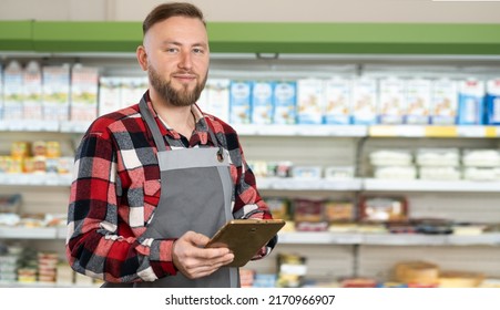 Portrait Of Sales Clerk Wearing Apron Using A Digital Tablet With Store Shelves On Background, Banner, Free Space