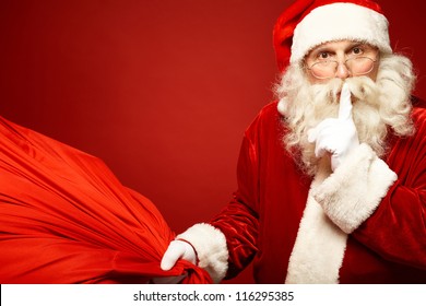 Portrait of Santa Claus with huge red sack keeping forefinger by his mouth and looking at camera - Powered by Shutterstock