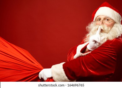 Portrait of Santa Claus with huge red sack keeping forefinger by his mouth and looking at camera