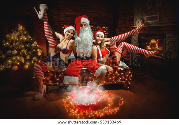 Portrait of Santa Claus sitting on the bed and steaming his legs around two sexy girls in Santa hats 