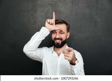 Portrait Of Sarcastic Man Showing Loser Sign On His Forehead And Pointing On Camera With Smile Mocking Or Humiliate Over Graphite Wall