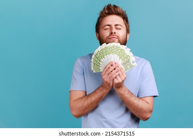 Portrait Of Satisfied Bearded Man Smelling Earned Euro Banknotes, Enjoying Success And Big Profit, Wealthy Life, Greedy For Money. Indoor Studio Shot Isolated On Blue Background.