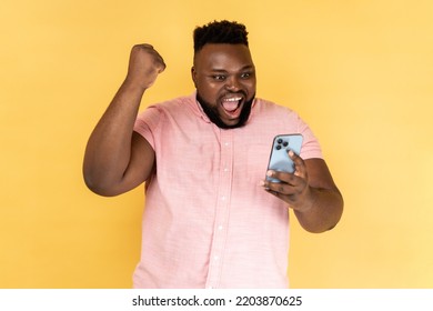 Portrait of satisfied bearded man wearing pink shirt standing with cell phone in hands and clenching fist, celebrating his victory, looks at screen. Indoor studio shot isolated on yellow background. - Shutterstock ID 2203870625