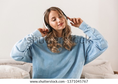 Portrait of satisfied blond girl 20s wearing headphones smiling and listening to music while sitting on sofa at home