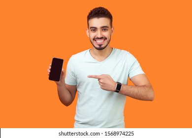 Portrait Of Satisfied Brunette Man With Beard In White T-shirt Pointing At Cellphone And Smiling At Camera, Recommending Gadget Or Mobile Application. Indoor Studio Shot Isolated On Orange Background