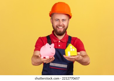 Portrait Of Satisfied Builder Man Wearing Blue Uniform And Protective Helmet Showing Piggy Bank And Paper House, Looking At Camera. Indoor Studio Shot Isolated On Yellow Background.