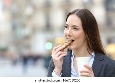 Portrait Of A Satisfied Business Woman Eating A Cereal Snack Bar Holding A Coffee Takeaway Cup On A City Street