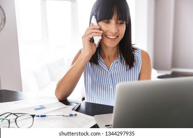 Portrait von zufriedener Geschäftsfrau der 40er in elegantem Kleidungsstück mit Smartphone und silbernem Laptop in hellem Raum