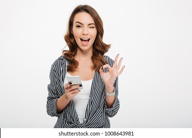 Portrait of a satisfied casual girl holding mobile phone and showing ok gesture isolated over white background - Powered by Shutterstock