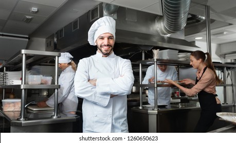 Portrait Of Satisfied Cheerful Positive  Smiling Chef On Restaurant Kitchen With Busy Professional Staff