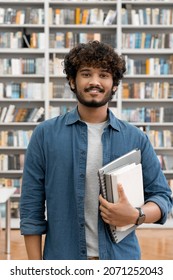 Portrait Of Satisfied Confident Smiling Young Indian Male Freelancer Or Student Holding Books Looking At Camera Standing In University Library. Research For Class Assignment And Exams Preparation