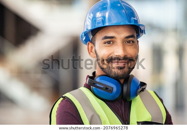 Portrait of satisfied construction site manager\
wearing safety vest and blue helmet with copy space. Young middle\
eastern architect watching construction site with confidence and\
looking at camera.