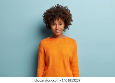 Portrait Of Satisfied Dark Skinned Female Model With Afro Haircut, Gentle Smile, Dressed In Casual Orange Jumper, Looks Straightly At Camera, Pose Over Blue Studio Wall For Making Photo. Ethnicity