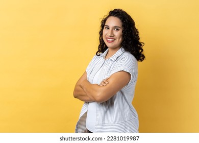 Portrait of satisfied delighted happy woman with dark wavy hair standing with folded hands and looking at camera with toothy smile. Indoor studio shot isolated on yellow background. - Shutterstock ID 2281019987