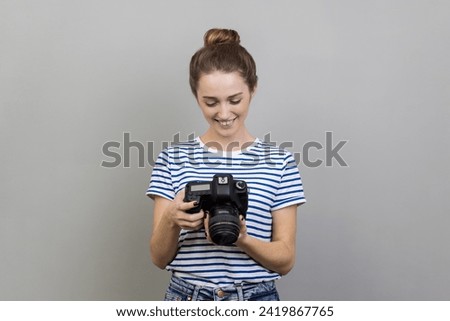 Portrait of satisfied delighted woman traveler or photographer wearing striped T-shirt holding and looking at photocamera, taking photo enjoying hobby. Indoor studio shot isolated on gray background.