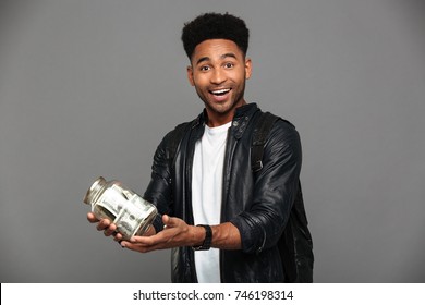 Portrait Of A Satisfied Excited Afro American Man In Leather Jacket Holding Jar Of Money Banknotes And Looking At Camera Isolated Over Gray Background