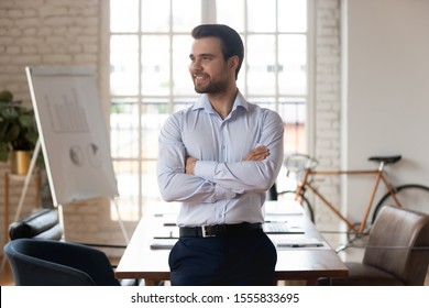 Portrait of satisfied millennial caucasian businessman posing with arms crossed standing inside of modern office boardroom looking away dreaming planning future successful projects, leadership concept