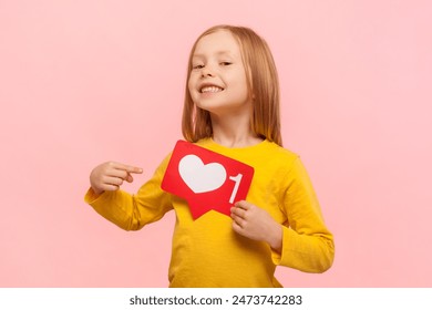 Portrait of satisfied proud blonde little girl holding pointing at blogger heart, looking at camera with smile, internet blogging, wearing yellow jumper. Indoor studio shot isolated on pink background - Powered by Shutterstock