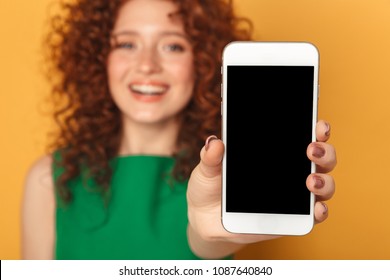 Portrait of a satisfied redhead woman in dress showing blank screen mobile phone isolated over yellow background