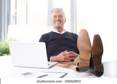 Portrait of satisfied senior businessman sitting at workplace in front of computer and looking at camera while relaxing. Old professional man feet on office desk. 