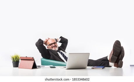 Portrait Of A Satisfied Senior Indian Asian Businessman Sitting At Workplace In Front Of The Computer And Looking At The Camera While Relaxing. Old Professional Man Feet On Office Desk