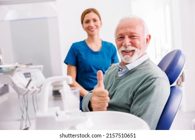 Portrait Of Satisfied Senior Male Patient Sitting At Dental Chair At Dentist Office Showing Thumbs Up And Smiling With Doctor In The Background