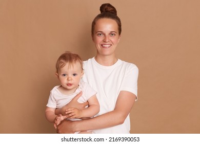 Portrait Of Satisfied Smiling Mother Holding Infant Baby Girl, Happy Family Having Fun Together, Adorable Kid In Mom's Arms, Mom And Child Isolated Over Brown Background.