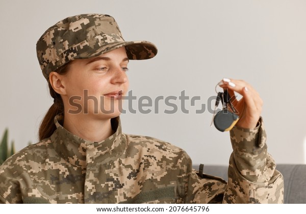 Portrait of satisfied woman\
soldier wearing camouflage uniform and hat, posing at home, holding\
key in hands, being happy to buy new purchase, house or\
car.