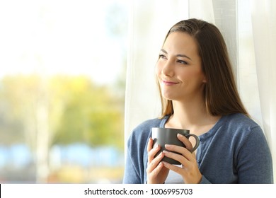 Portrait of a satisfied woman thinking looking at side holding a coffee mug beside a window at home - Shutterstock ID 1006995703