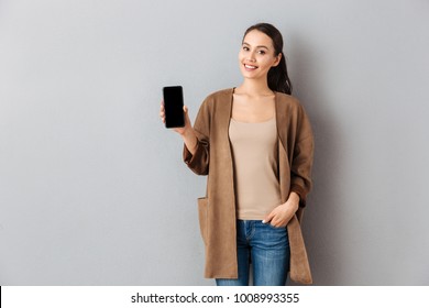 Portrait of a satisfied young asian woman showing blank screen mobile phone while standing and looking at camera over gray background