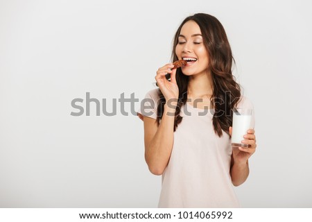 Portrait of a satisfied young asian woman eating cookie and holding glass of milk isolated over white background