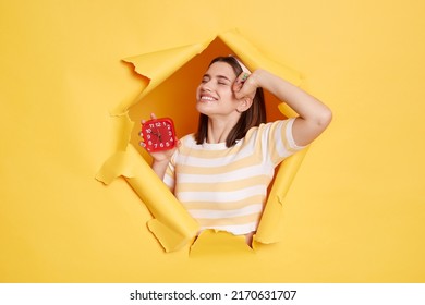 Portrait Of Satisfied Young Caucasian Woman Wearing Striped Shirt And Hair Ban D Posing In Yellow Paper Hole, Standing And Stretching Body After Sleeping, Wakes Up In Good Mood, Holding Alarm Clock.