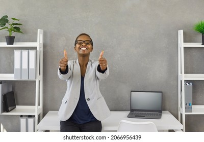 Portrait Of Satisfied Young Lady At Work. Happy Excited Black Business Woman Standing By Office Desk With Laptop Computer, Looking At Camera, Smiling And Giving Thumbs Up With Both Hands