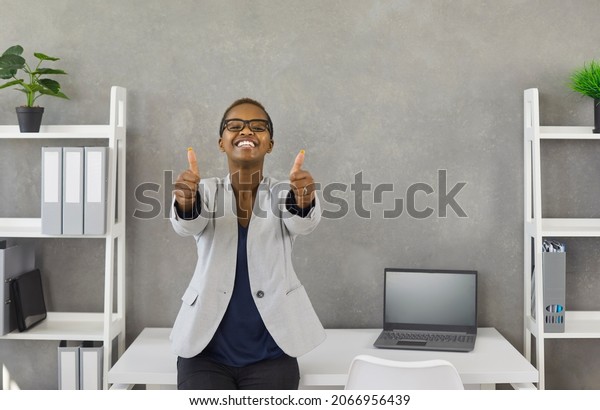 Portrait of\
satisfied young lady at work. Happy excited black business woman\
standing by office desk with laptop computer, looking at camera,\
smiling and giving thumbs up with both\
hands