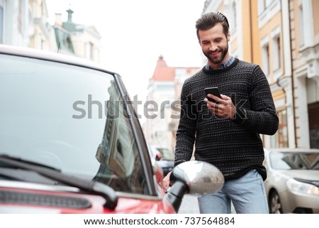 Portrait of a satisfied young man in sweater using mobile phone while standing at his car outdoors