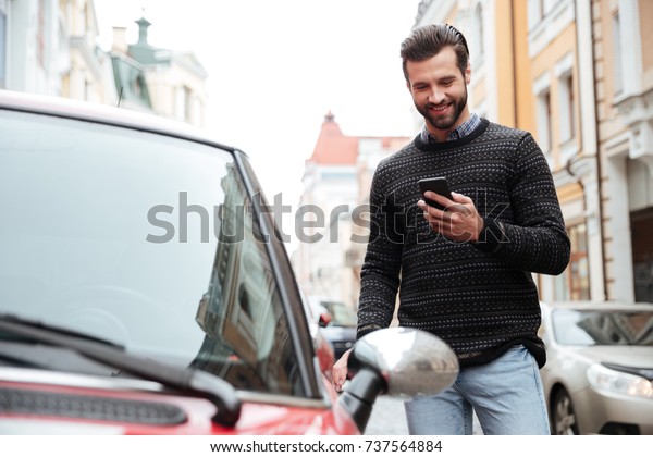 Portrait of a satisfied\
young man in sweater using mobile phone while standing at his car\
outdoors