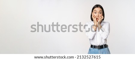 Portrait of scared asian woman watching smth scary, biting fingers on hands and looking at camera frightened, standing over white background