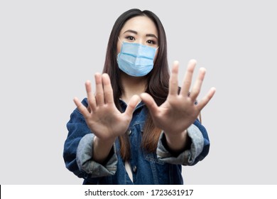 Portrait Of Scared Beautiful Asian Young Woman With Surgical Medical Mask In Blue Denim Jacket Standing With Hand Blocking And Looking At Camera. Indoor Studio Shot, Isolated On Grey Background