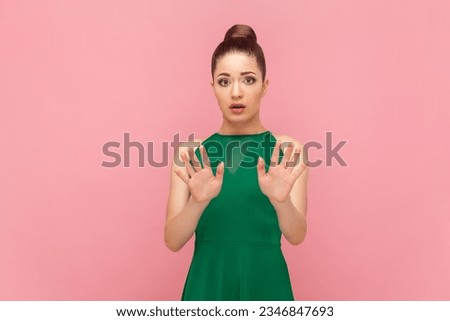 Portrait of scared frighten woman with bun hairstyle standing showing palms to camera, demonstrates stop sign, asking to stop it, wearing green dress. Indoor studio shot isolated on pink background.