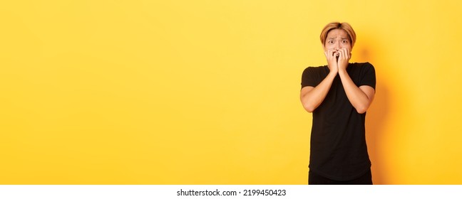 Portrait Of Scared Insecure Asian Blond Guy, Holding Hands Over Mouth Horrified, Looking Frightened, Yellow Background