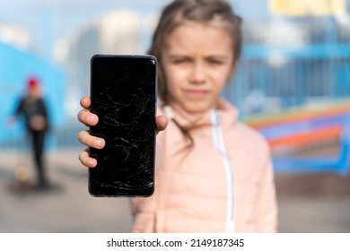 Portrait Of  Scared Kid Girl With Broken Mobile Phone At Skatepark. Sad Child Broke Screen Of Mobile Phone. Little Girl In Desperation Is Holding Phone With Cracked Screen At Day Time. 