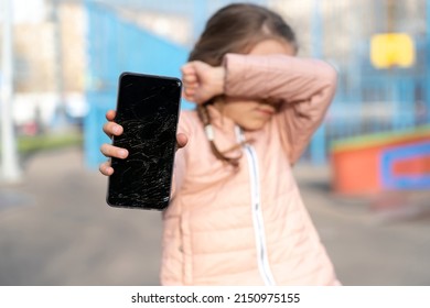 Portrait Of  Scared Kid Girl With Broken Mobile Phone At Skatepark. Sad Child Broke Screen Of Mobile Phone. Little Girl In Desperation Is Holding Phone With Cracked Screen At Day Time. 