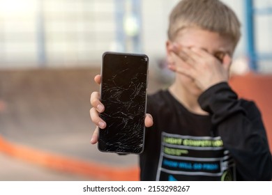 Portrait Of  Scared Kid Girl With Broken Mobile Phone At Skatepark. Sad Child Broke Screen Of Mobile Phone. Teen Boy In Desperation Is Holding Phone With Cracked Screen At Day Time. 