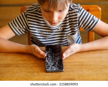 Portrait Of Scared Kid Girl With Broken Mobile Phone On Table. Sad Child Broke Screen Of Mobile Phone. Teen Boy In Desperation Is Holding Phone With Cracked Screen At Day Time.