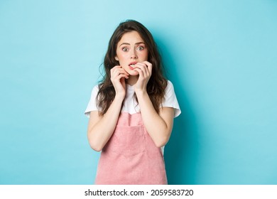 Portrait of scared and startled brunette girl biting fingernails, staring at camera shocked and worried, hear terrible news, standing in stupor against blue background