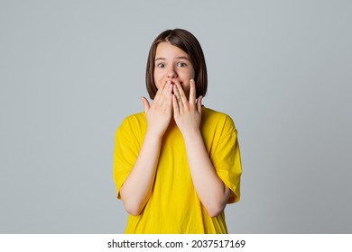 Portrait Of A Scared Teen Girl Looking At Camera With Wide Opened Eyes, Covering Mouth Her Hands, Stand Over Light Grey Background