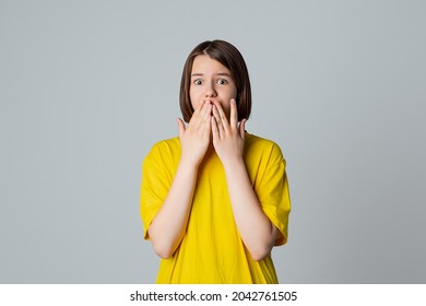 Portrait Of A Scared Teen Girl Looking At Camera With Wide Opened Eyes, Covering Mouth Her Hands, Stand Over Light Grey Background