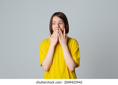 Portrait Of A Scared Teen Girl, Looking Away With Wide Opened Eyes, Covering Mouth Her Hands, Stand Over Light Grey Background