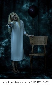 Portrait of a scary pale girl  with black balloon next to the chair on grunge background. Zombie, halloween.