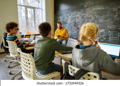 Portrait Of Schoolgirl And Schoolboys Using Laptops During A Lesson In Modern Smart School. Male Teacher In The Background. STEM Disciplines